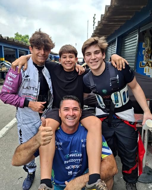Zé do Bode ao lado de três jovens pilotos de kart de macacão, no box